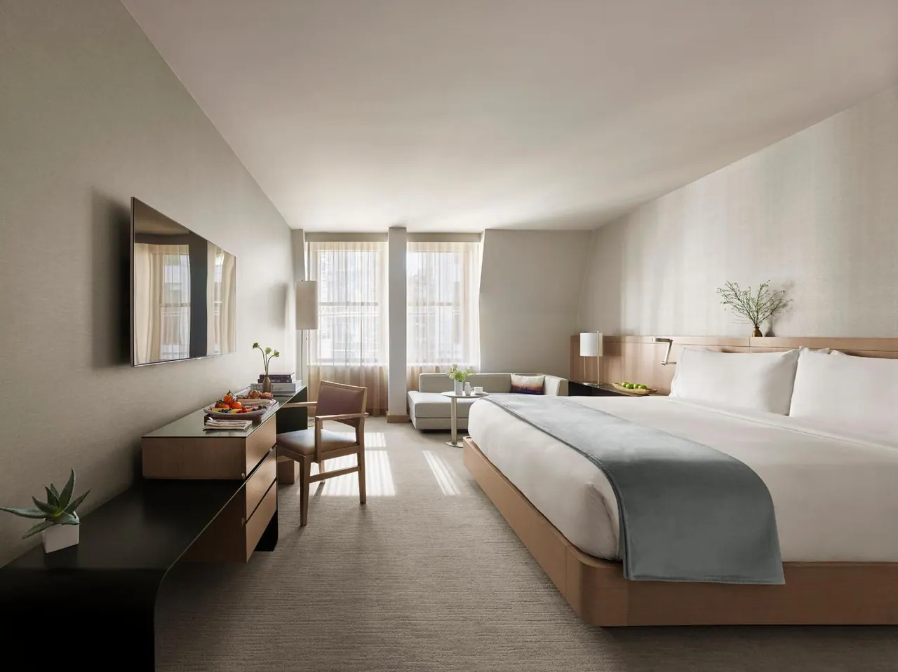 A bedroom with a king bed, wall-mounted television, wooden desk with chair, and a grey sofa by tall windows.