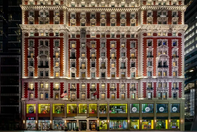 Night view of the ornate red-brick and stone facade of the Knickerbocker Hotel, with lit windows and ground-floor shop fronts.