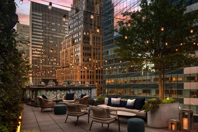 A rooftop terrace with wicker armchairs, a sofa with navy cushions, a tree in a planter and string lights, with tall city buildings behind at dusk.