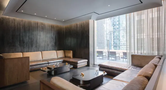 A lounge with leather sectional sofas, two low coffee tables and floor-to-ceiling windows looking onto a terrace and high-rise buildings.