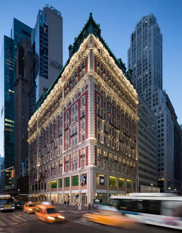 Exterior of an illuminated corner building with ornate cornices and a red brick facade at dusk, with taxis and a bus passing on the street below.