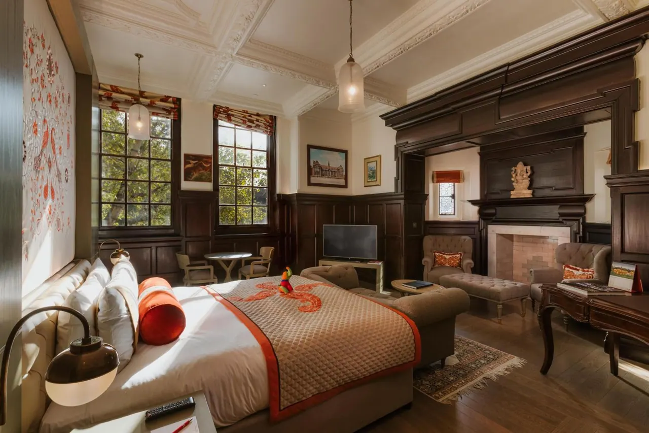 A large hotel bedroom with a king bed, a seating area, a fireplace, dark wood panelling and tall windows looking onto trees.