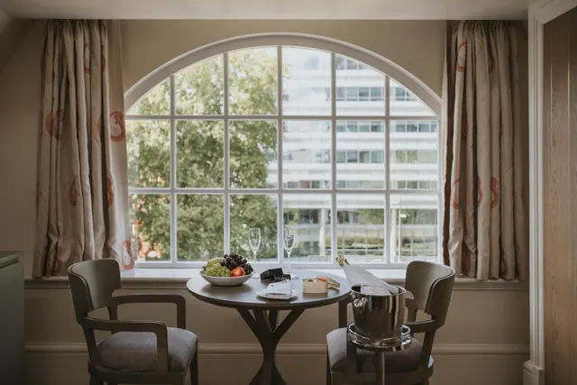 A round table by an arched window holds a fruit bowl, two wine glasses, a cheese plate and a champagne bucket, with two upholstered chairs beside it.