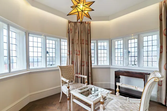 A bay-windowed room with a star-shaped pendant light, two upholstered chairs, a glass-topped table and a dark wood console beneath tall windows.