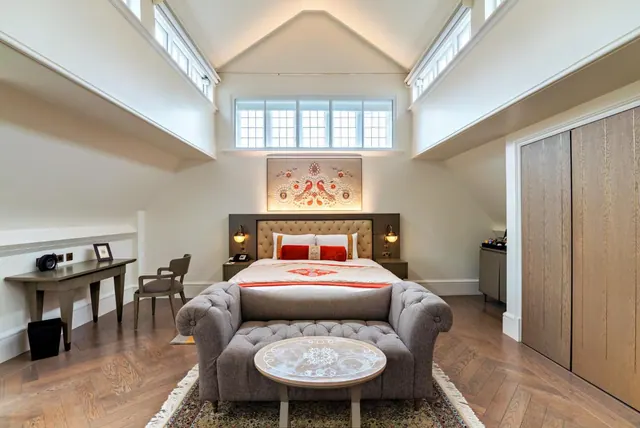 A bedroom with a double bed under a sloped ceiling, a tufted grey sofa at the foot of the bed, a writing desk and chair on the left, and a wardrobe on the right.