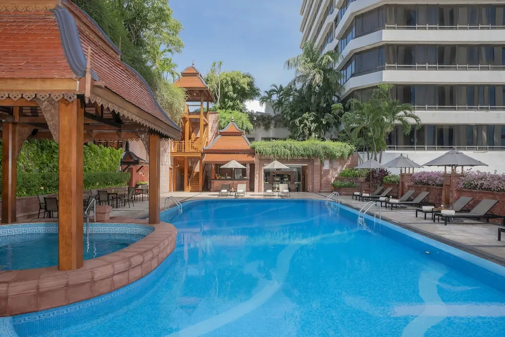 An outdoor pool with a large lap pool and a small round pool on the left, surrounded by sun loungers and shade umbrellas beside a Thai-style pavilion and a tall balconied building.