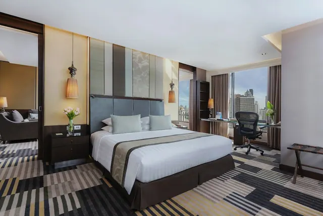 A bedroom with a made king bed, a desk and chair by a large window, and a view of city high-rise buildings, with a seating area visible through the open doorway on the left.