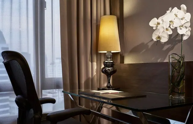 A glass desk with a black mesh office chair, a lit table lamp, and a white orchid in a glass vase sits beside a window with blurred city buildings outside.