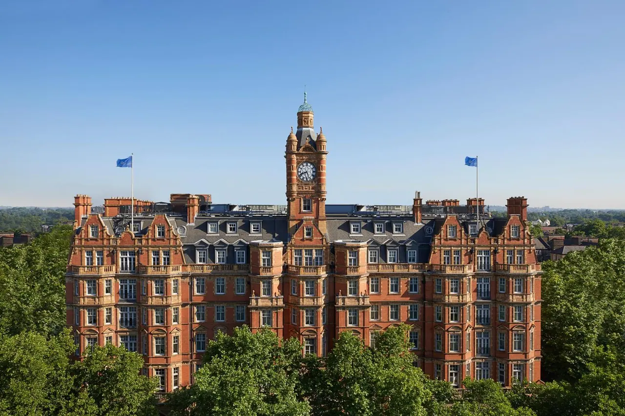 Elevated exterior view of a red brick Victorian building with a clock tower and two blue flags, surrounded by trees under a clear sky.