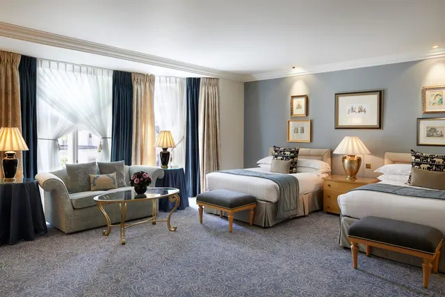 A hotel bedroom with two double beds with grey throws, a sofa and coffee table by the windows, and framed prints on a blue-grey wall.
