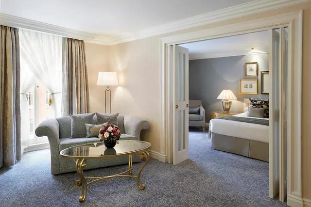 A living area with a grey sofa, a glass-topped coffee table holding a flower arrangement, and an open door into a bedroom with a double bed.