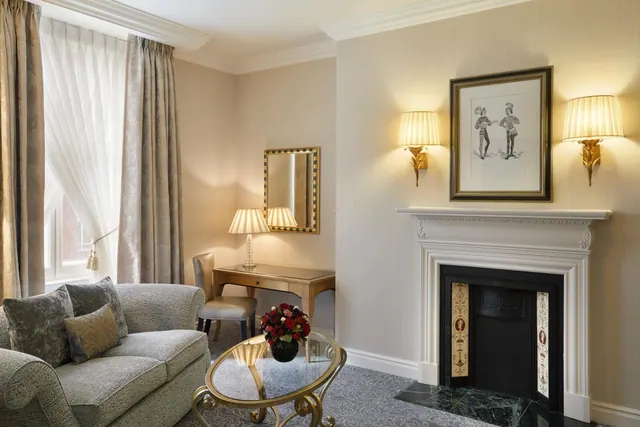 A living area with a grey upholstered sofa, a glass-topped gold coffee table, a white fireplace with a black insert, and a small writing desk beside a window with sheer curtains.