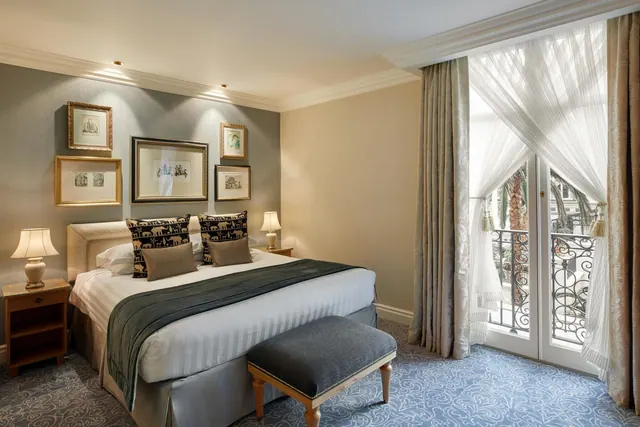 A bedroom with a double bed, framed pictures on the wall, bedside lamps, and French doors opening onto a balcony with a wrought iron railing.