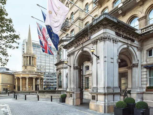 The stone front entrance of The Langham hotel with flags hanging above, and a domed church with a spire across the street on the left.