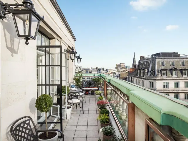 A long rooftop terrace with potted plants, metal chairs and a cushioned bench, a green awning, and views over surrounding buildings and a church spire.