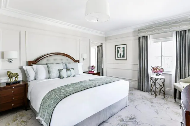 A bedroom with a king bed dressed in white and grey-green linens, wood nightstands and wall sconces on either side, grey curtains, and a window on the right.