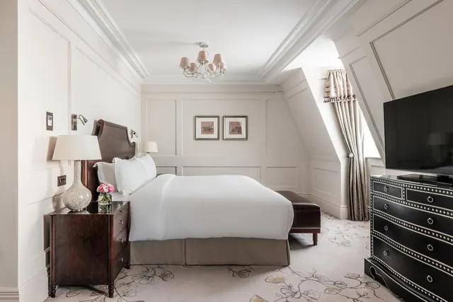 A bedroom with a made double bed, a bedside table with a lamp, a flat-screen television on a black dresser, and curtains by a window.