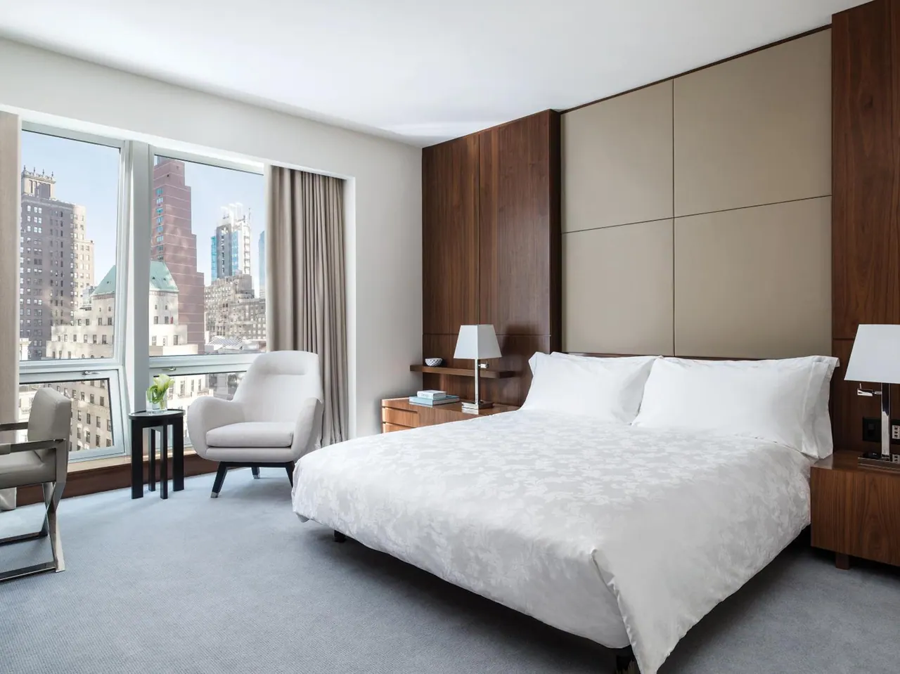 A bedroom with a king bed, an armchair beside a large window looking out over city buildings, and a wood-panelled headboard wall.