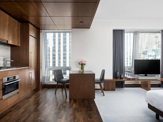A studio hotel room with a wood kitchenette, a desk with two chairs, a wall-mounted TV and a bed at the right edge, with city buildings visible through the windows.