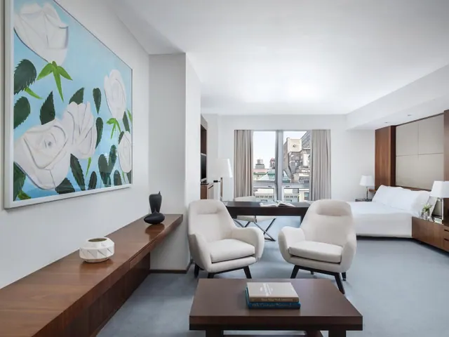 A bright hotel room with a large painting, two upholstered armchairs and a coffee table, with a bed and desk near the window looking out over city buildings.