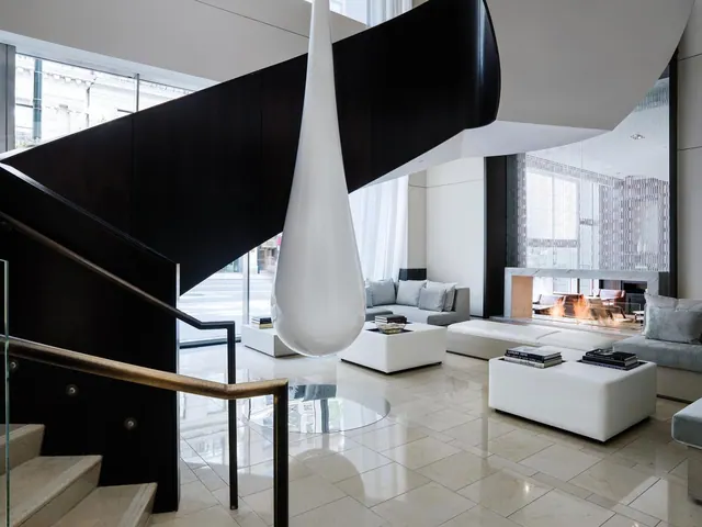 A lobby with a black staircase, a suspended white teardrop sculpture, white ottomans and sofas on a stone floor, a fireplace in the background, and a street visible through the windows.