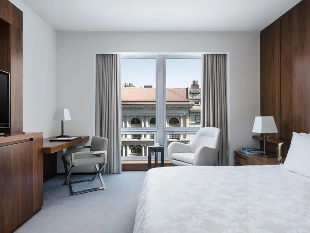 A hotel bedroom with a king bed, a desk, an armchair and a window looking out onto a stone building with a red tiled roof.