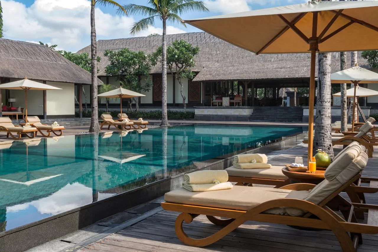 A large swimming pool with teak sun loungers, folded towels and parasol umbrellas on a timber deck, with thatched-roof pavilions and palm trees behind.