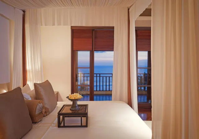 A bedroom with a canopy bed and sheer curtains, with sliding glass doors opening onto a balcony with a view of the sea at dusk.
