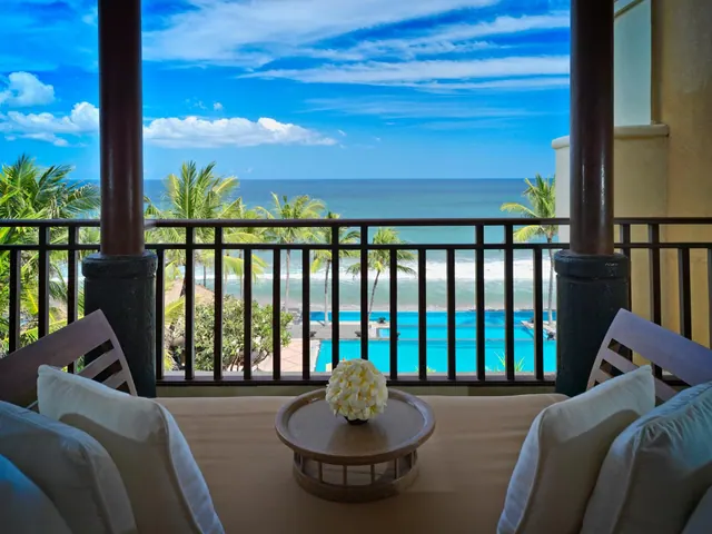 A covered balcony with a cushioned daybed and chairs and a small table with white flowers, overlooking a swimming pool, palm trees and the ocean.