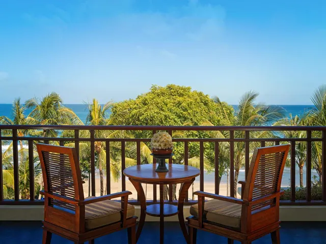 A wooden balcony with two armchairs and a small round table overlooks palm trees, a sandy beach and the sea.