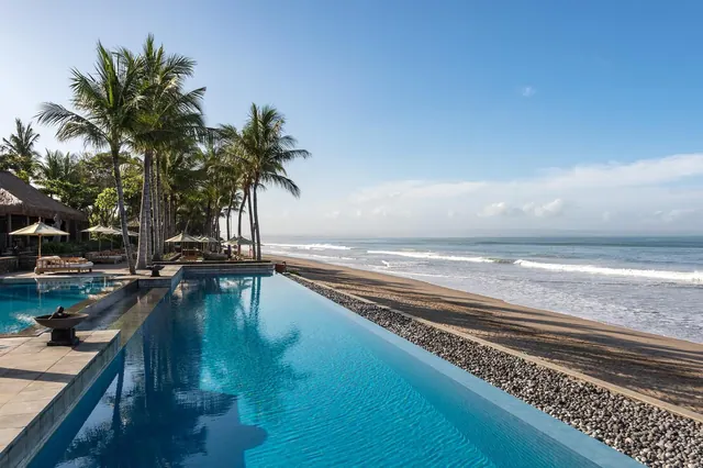 An infinity-edge swimming pool runs alongside a sandy beach with palm trees and the ocean beyond.