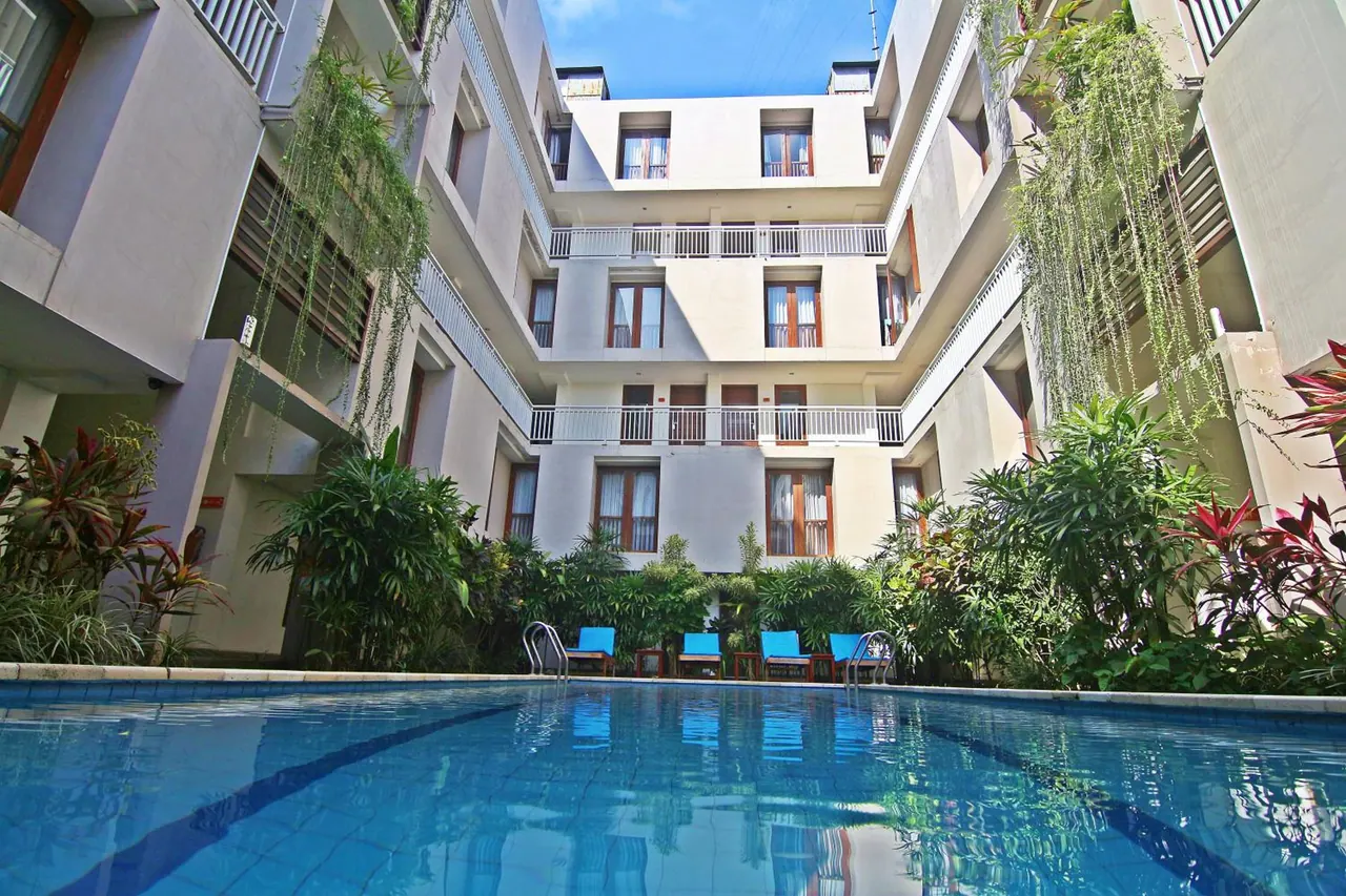 A swimming pool in a central courtyard with blue lounge chairs and tropical plants, surrounded by a multi-storey hotel building with balconies.