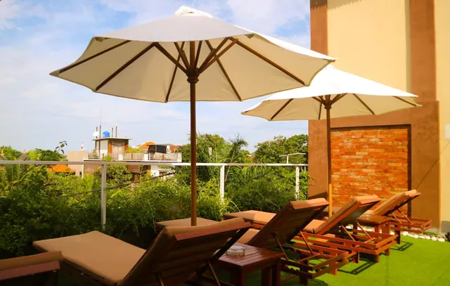 A terrace with beige parasol umbrellas, wooden sun loungers and side tables on artificial grass, with a brick wall and tropical greenery beyond the railing.