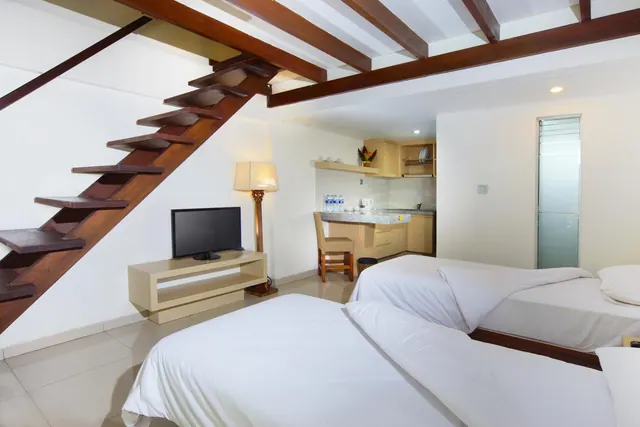 A bedroom with two white beds, a wooden staircase to a loft, a flat-screen television on a low stand, and a small kitchenette in the background.