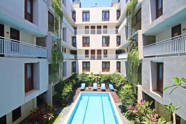 A long rectangular swimming pool in a courtyard with blue sun loungers, surrounded by multi-storey building balconies and tropical plants.