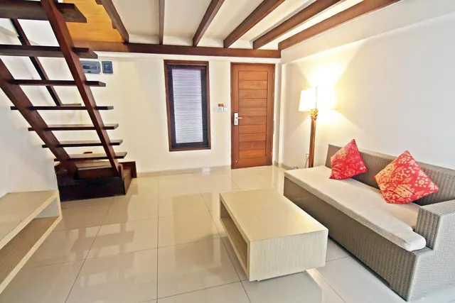 A living area with a wicker sofa and red patterned cushions, a low wooden coffee table, an open wooden staircase to a loft, and a wooden front door.