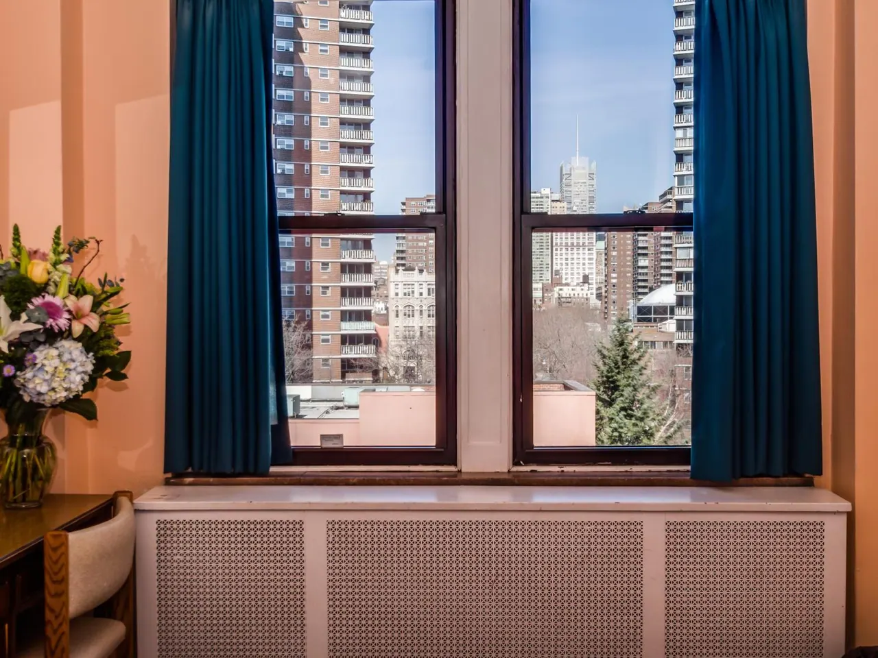 A pair of windows with dark teal curtains looks out over high-rise residential buildings and a distant spired tower, with a flower arrangement and a chair at the left edge.