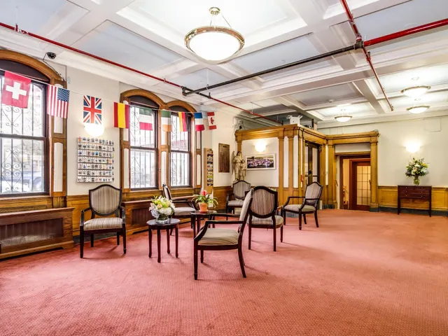 A lobby lounge with red carpet, striped armchairs and small side tables, national flags hung across tall arched windows, and a coffered ceiling.