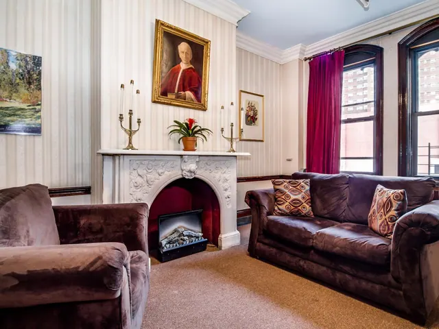 A living room with a marble fireplace, a framed portrait above the mantel, a brown sofa and armchair, and arched windows looking onto a brick building.