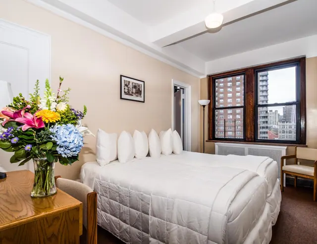 A bedroom with a double bed, a vase of flowers on a wooden side table, and a window looking out over brick high-rise buildings.