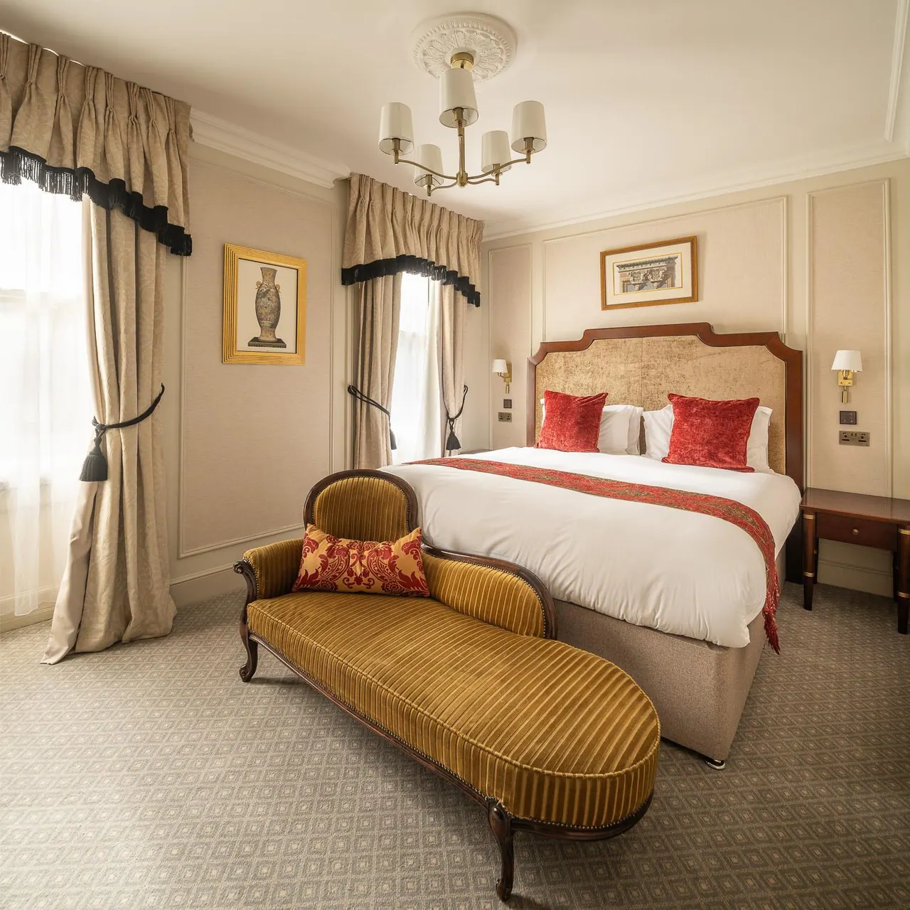 A bedroom with a made double bed, red cushions, a gold striped chaise longue, and tall curtains beside a window.