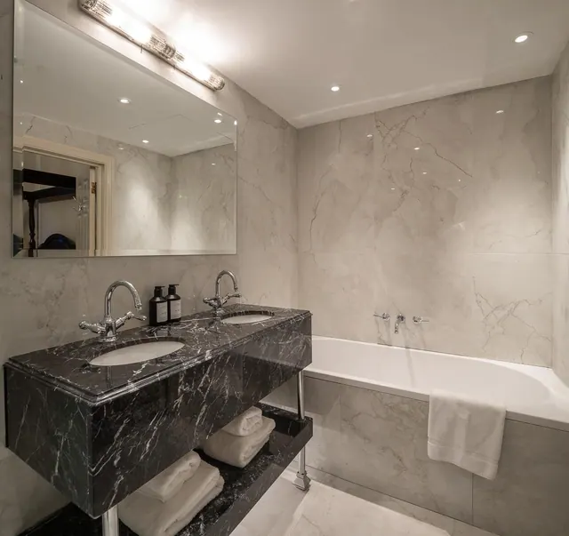A bathroom with a marble double vanity under a wall-mounted light, a built-in bathtub with wall-mounted taps, and folded white towels on a dark marble shelf.