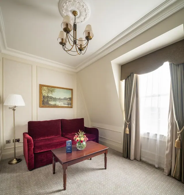 A furnished sitting area with a red velvet sofa, a wooden coffee table with a flower vase, a floor lamp, a chandelier and a framed landscape painting beside a curtained window.