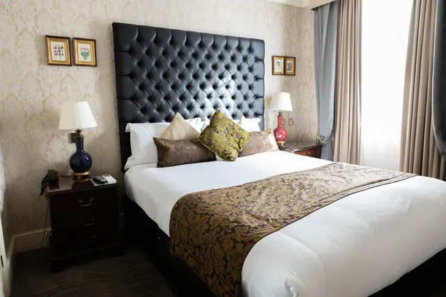 A made double bed with a tall tufted black leather headboard, a patterned bed runner, and bedside lamps sits beneath framed botanical prints on a damask-patterned wall.