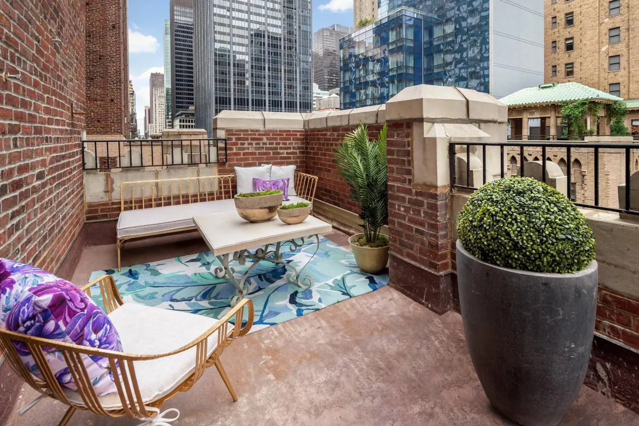 A brick-walled rooftop terrace with a bench, a patterned rug, a table, a rattan chair and potted plants, with tall office towers visible behind.