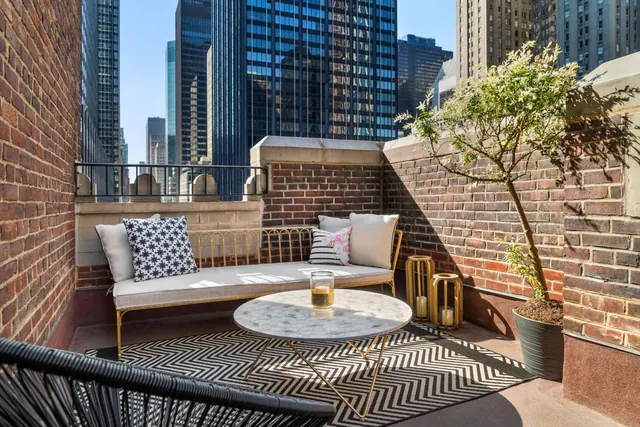 A brick-walled terrace with a gold daybed, a round marble table, a patterned rug and a potted tree, with tall city towers visible beyond the wall.