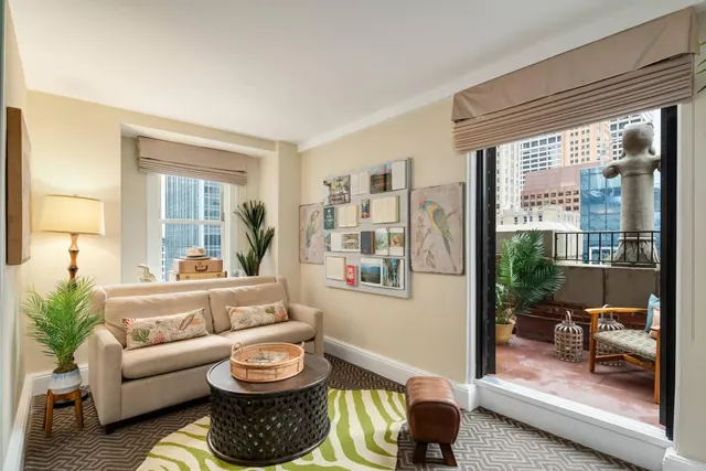 A small living area with a beige sofa, table lamp and wall of framed pictures, with a glass door opening onto a balcony overlooking high-rise buildings.