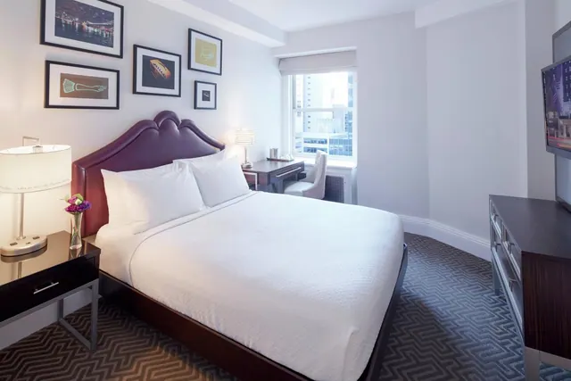 A bedroom with a king bed, a desk by the window, a wall-mounted television and framed artwork on the wall.