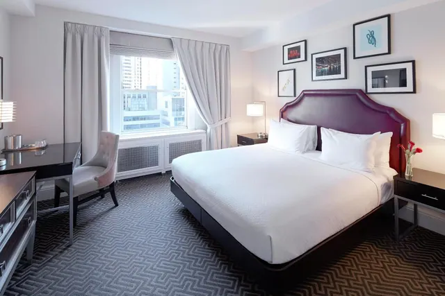 A bedroom with a made-up king bed under a dark upholstered headboard, a desk and upholstered chair by the window, and high-rise buildings visible outside.
