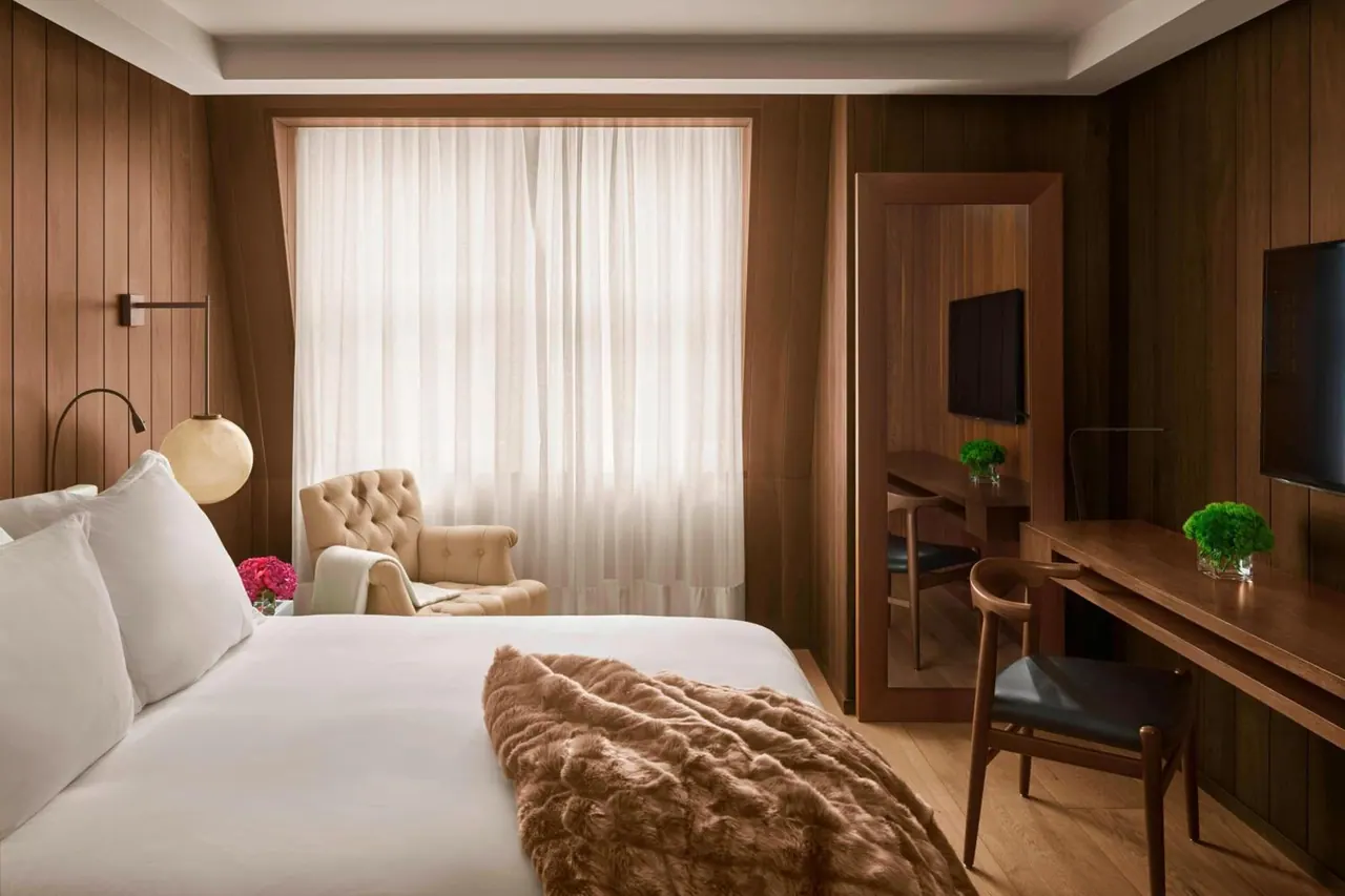 A bedroom with a made white bed, a tufted armchair by a sheer-curtained window, a wooden desk with a chair, and a wall-mounted television.